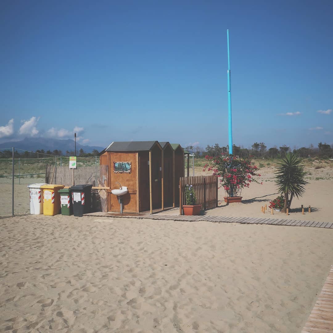 Scena di spiaggia in Toscana con bidoni per la raccolta rifiuti e strutture balneari, tra cui bagni da spiaggia e cabine in legno. Alcuni vasi con vegetazione mediterranea aggiungono un tocco di verde all’ambiente. L’atmosfera rilassata richiama la tipica villeggiatura estiva italiana.